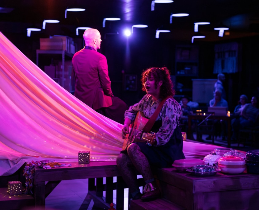 A dimly lit scene with purple and pink lighting, featuring a woman seated on a wooden platform playing an acoustic guitar. Another individual in a suit sits nearby, facing away. Decorative items and casserole dishes are arranged around the platform, while the audience observes the performance.