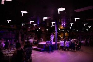 A dimly lit room, featuring a central stage where one person speaks into a microphone and another plays an acoustic guitar. The audience is seated around the stage at tables and chairs.