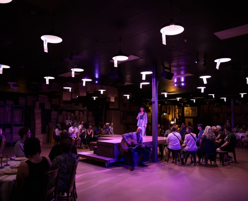 A dimly lit room, featuring a central stage where one person speaks into a microphone and another plays an acoustic guitar. The audience is seated around the stage at tables and chairs.
