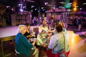 Five people sit around a small table sharing tea in a room filled with shelves stacked with cardboard boxes. The space is illuminated with colorful lights.