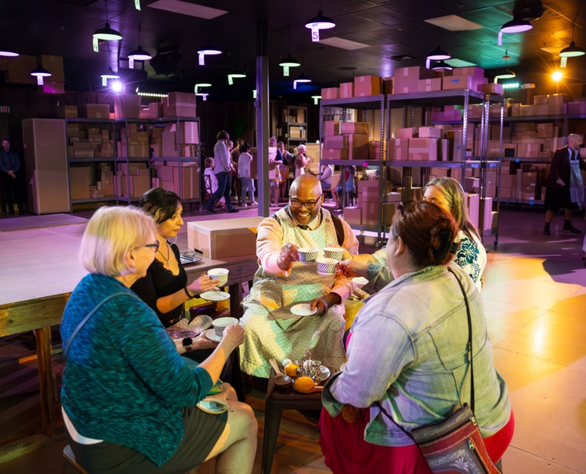 Five people sit around a small table sharing tea in a room filled with shelves stacked with cardboard boxes. The space is illuminated with colorful lights.