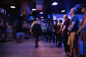 Five people sit around a small table sharing tea in a room filled with shelves stacked with cardboard boxes. The space is illuminated with colorful lights.