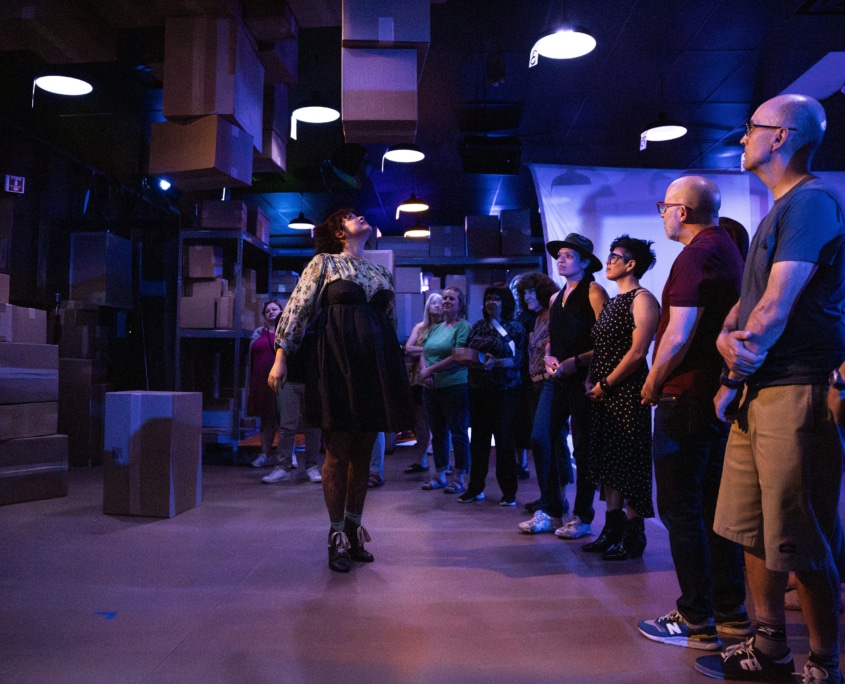 Five people sit around a small table sharing tea in a room filled with shelves stacked with cardboard boxes. The space is illuminated with colorful lights.