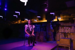 A person sits on a chair in a dimly lit room with purple and yellow lighting. The background features shelves stacked with cardboard boxes. Lights hang from the ceiling, illuminating the space.
