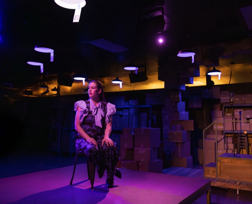 A person sits on a chair in a dimly lit room with purple and yellow lighting. The background features shelves stacked with cardboard boxes. Lights hang from the ceiling, illuminating the space.