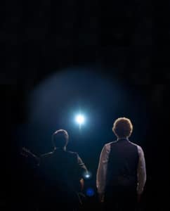 Back view of two performers under a bright spotlight, one holding a guitar.