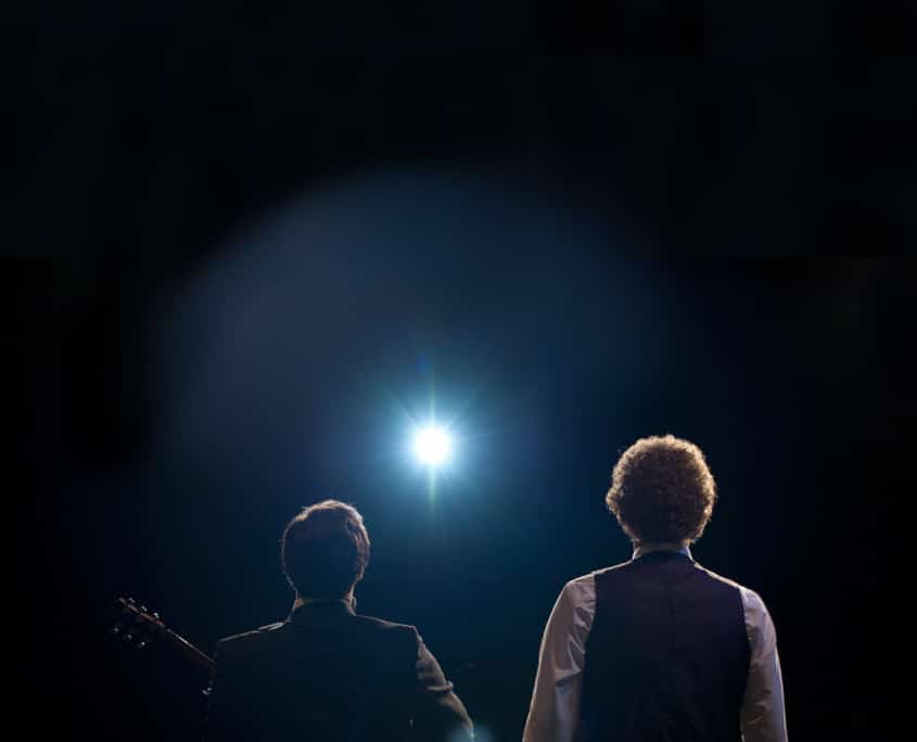 Back view of two performers under a bright spotlight, one holding a guitar.