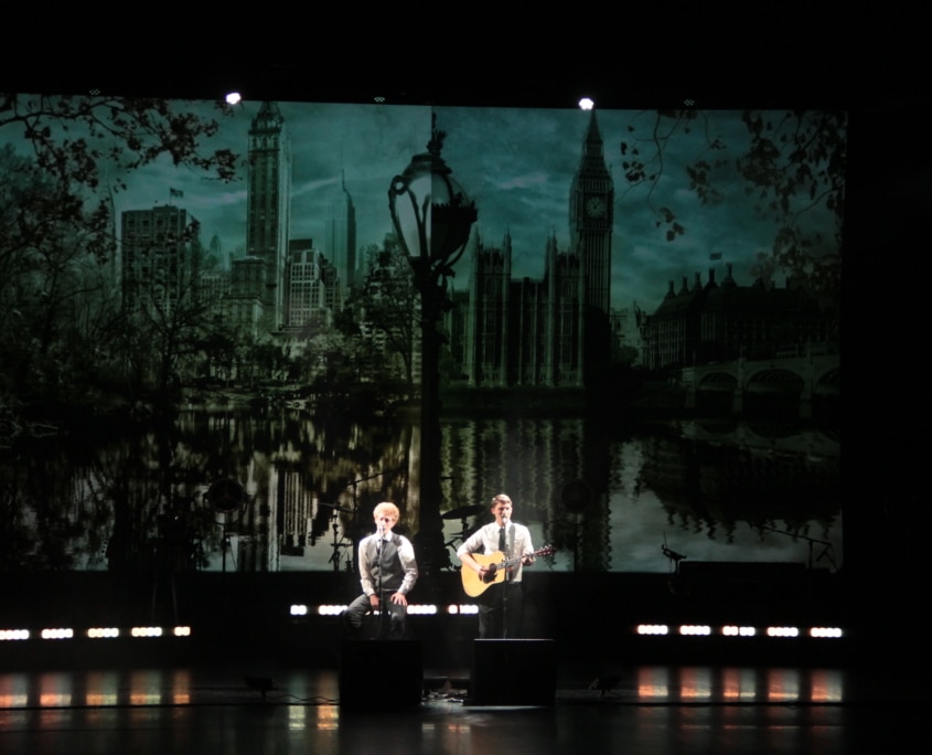 Two musicians perform on stage with a London cityscape backdrop, one playing guitar.