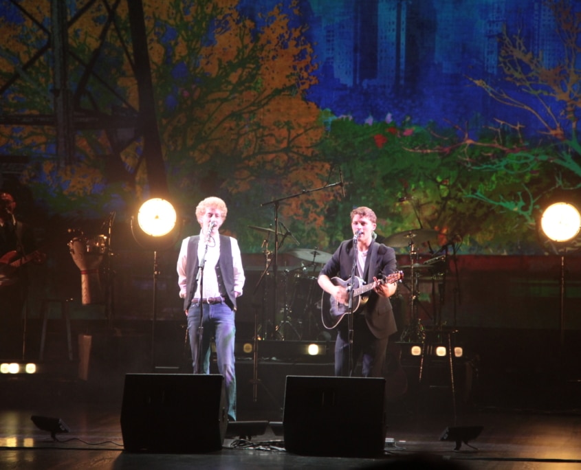 Two musicians perform on stage with a London cityscape backdrop, one playing guitar.2