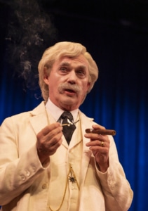 Mark Twain dressed in a cream-colored three-piece suit with a black tie and gold pocket watch chain is holding a lit match in one hand and a cigar in the other. Wisps of smoke rise in the air against a dark blue curtain backdrop.