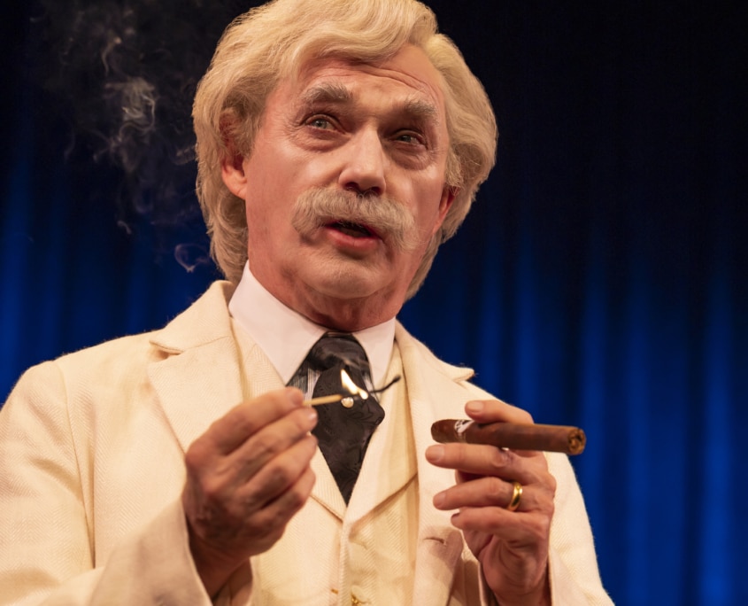 Mark Twain dressed in a cream-colored three-piece suit with a black tie and gold pocket watch chain is holding a lit match in one hand and a cigar in the other. Wisps of smoke rise in the air against a dark blue curtain backdrop.