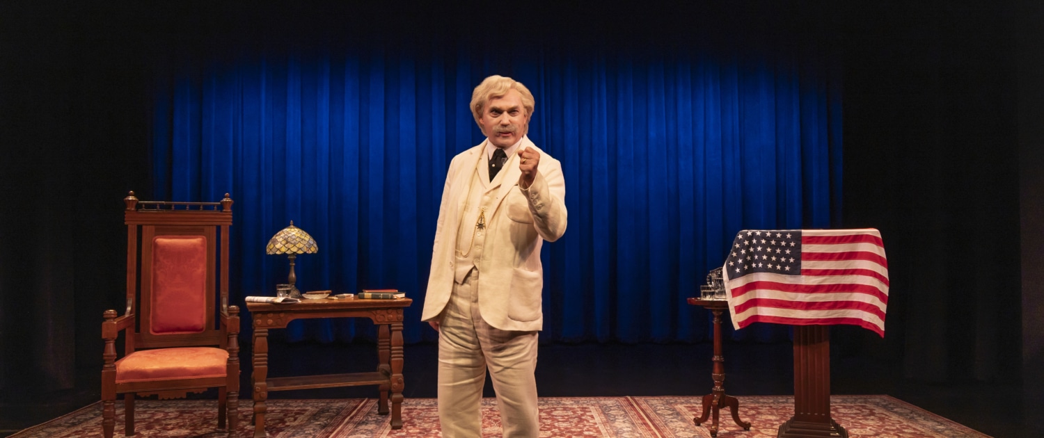 A person dressed as Mark Twain in a cream-colored suit stands center stage on a patterned rug, holding a cigar. The stage features a large blue curtain, a wooden desk with a lamp and books on the left, and a podium draped with an American flag on the right.