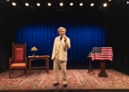 A person dressed as Mark Twain in a cream-colored suit stands center stage on a patterned rug, holding a cigar. The stage features a large blue curtain, a wooden desk with a lamp and books on the left, and a podium draped with an American flag on the right.