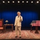 A person dressed as Mark Twain in a cream-colored suit stands center stage on a patterned rug, holding a cigar. The stage features a large blue curtain, a wooden desk with a lamp and books on the left, and a podium draped with an American flag on the right.