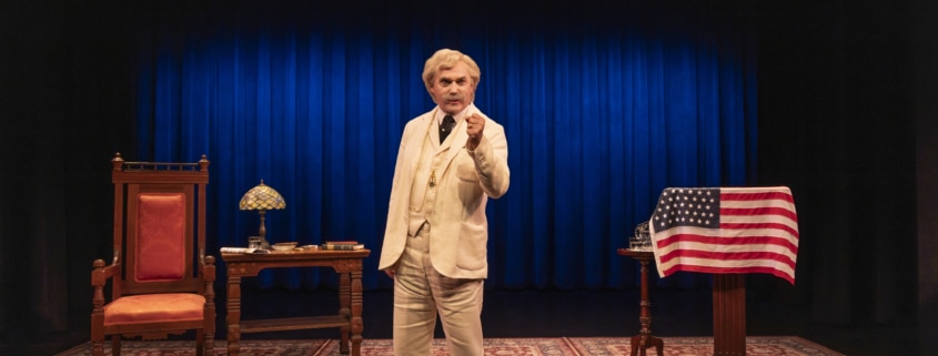 A person dressed as Mark Twain in a cream-colored suit stands center stage on a patterned rug, holding a cigar. The stage features a large blue curtain, a wooden desk with a lamp and books on the left, and a podium draped with an American flag on the right.