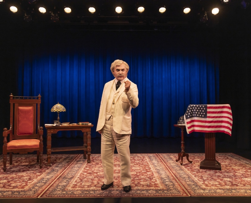A person dressed as Mark Twain in a cream-colored suit stands center stage on a patterned rug, holding a cigar. The stage features a large blue curtain, a wooden desk with a lamp and books on the left, and a podium draped with an American flag on the right.