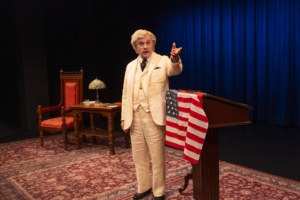 A person dressed as Mark Twain in a cream-colored suit stands beside a wooden podium draped with an American flag, gesturing with one hand. Behind them is a wooden desk with a decorative lamp and books, set against a deep blue curtain backdrop.