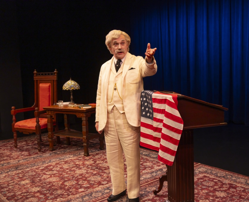 A person dressed as Mark Twain in a cream-colored suit stands beside a wooden podium draped with an American flag, gesturing with one hand. Behind them is a wooden desk with a decorative lamp and books, set against a deep blue curtain backdrop.