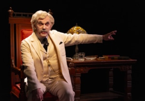 A person dressed as Mark Twain in a cream-colored suit sits in an ornate wooden chair at a desk with a stained-glass lamp, books, and a glass of water. The person extends one arm outward, pointing toward something off-frame, with a dark background emphasizing the warm lighting on the desk.