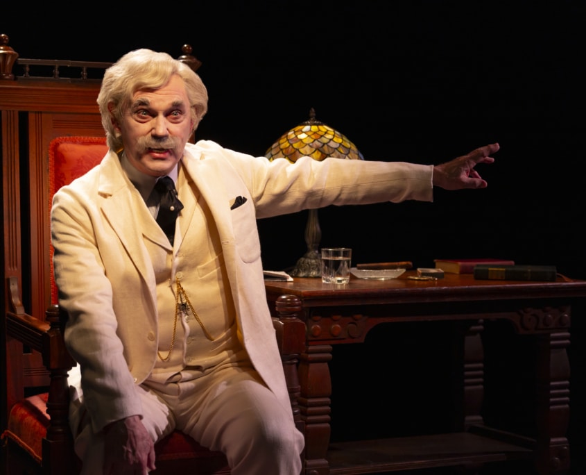 A person dressed as Mark Twain in a cream-colored suit sits in an ornate wooden chair at a desk with a stained-glass lamp, books, and a glass of water. The person extends one arm outward, pointing toward something off-frame, with a dark background emphasizing the warm lighting on the desk.
