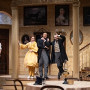 Denver Center Theatre Company world premiere of The Suffragette's Murder featuring four actors on a Victorian-era set with ornate wallpaper, framed portraits, and a staircase; one actor in a yellow dress, others in period suits and coats.