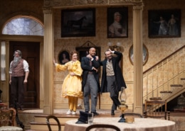 Denver Center Theatre Company world premiere of The Suffragette's Murder featuring four actors on a Victorian-era set with ornate wallpaper, framed portraits, and a staircase; one actor in a yellow dress, others in period suits and coats.