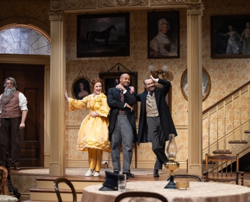 Denver Center Theatre Company world premiere of The Suffragette's Murder featuring four actors on a Victorian-era set with ornate wallpaper, framed portraits, and a staircase; one actor in a yellow dress, others in period suits and coats.