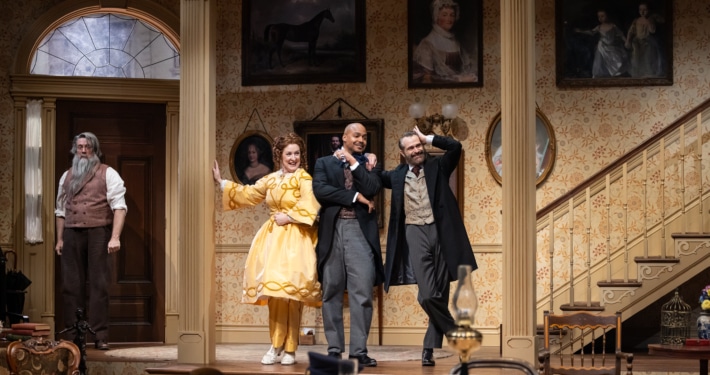 Denver Center Theatre Company world premiere of The Suffragette's Murder featuring four actors on a Victorian-era set with ornate wallpaper, framed portraits, and a staircase; one actor in a yellow dress, others in period suits and coats.