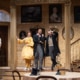 Denver Center Theatre Company world premiere of The Suffragette's Murder featuring four actors on a Victorian-era set with ornate wallpaper, framed portraits, and a staircase; one actor in a yellow dress, others in period suits and coats.