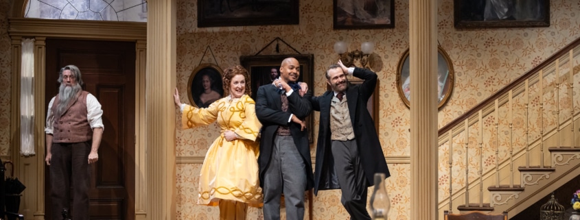 Denver Center Theatre Company world premiere of The Suffragette's Murder featuring four actors on a Victorian-era set with ornate wallpaper, framed portraits, and a staircase; one actor in a yellow dress, others in period suits and coats.