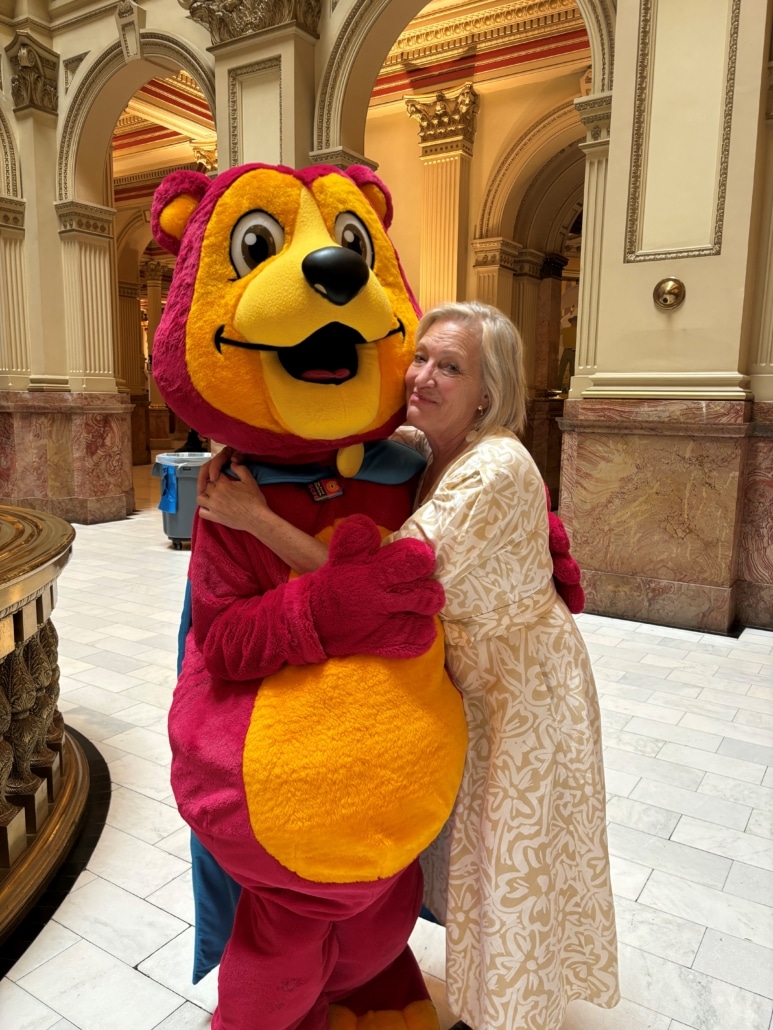 A person in a white dress hugs a large, colorful bear mascot inside an ornate building with arched doorways and detailed columns.