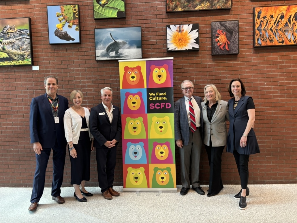 Group of six people standing in front of a colorful banner with bear faces and the text 'We Fund Culture. SCFD' against a brick wall adorned with various nature photographs.