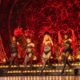 Alt tag: Four performers in elaborate costumes with feathered headdresses and fishnet stockings dance on a stage with an ornate, red and gold backdrop.
