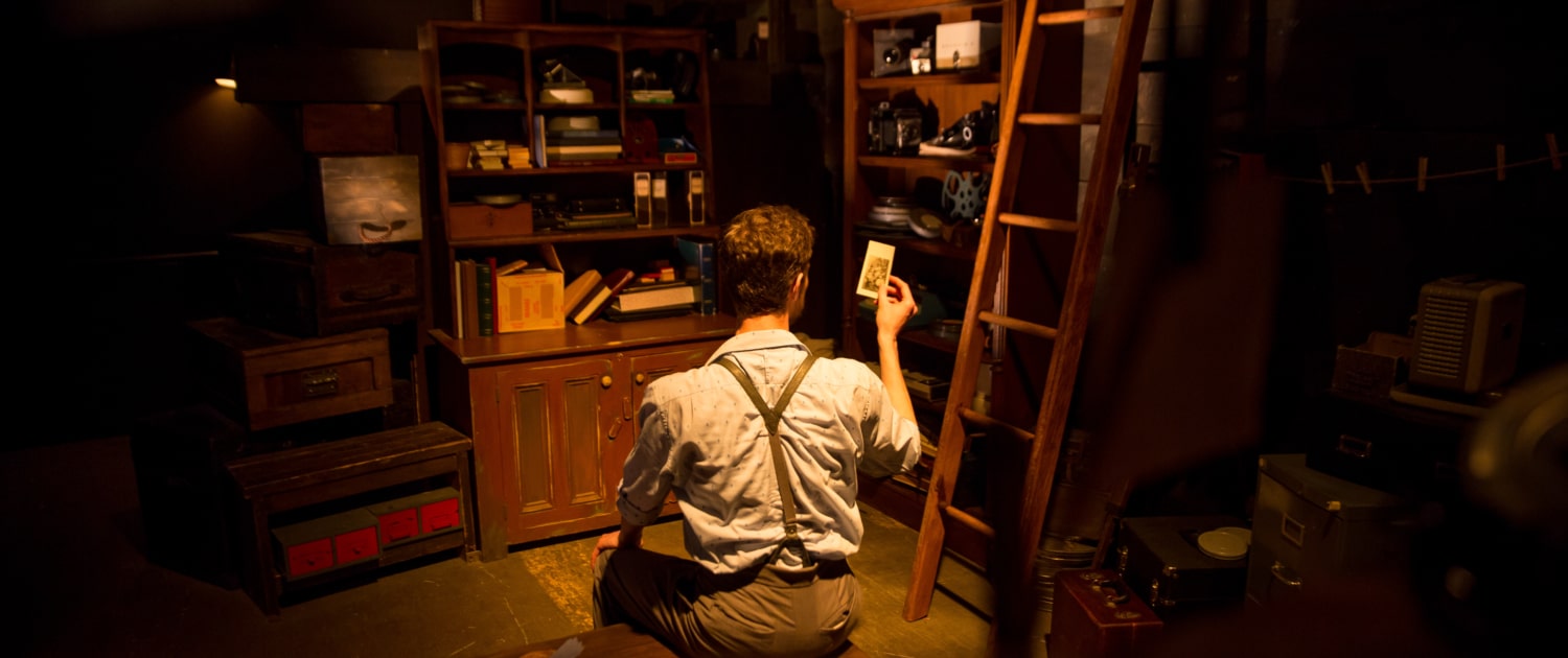 A person sitting on the floor in a dimly lit room, holding a photograph while gazing at shelves filled with assorted objects such as books and boxes. A wooden ladder leans against the shelves, contributing to the room’s rustic and contemplative atmosphere. 
