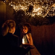 Two people standing beneath a canopy of glowing string lights. One person faces the camera, while the other stands with their back to the camera. The background includes foliage and a wooden fence, contributing to a warm, intimate atmosphere.