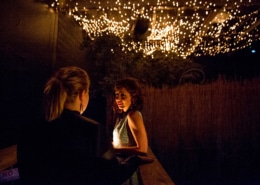 Two people standing beneath a canopy of glowing string lights. One person faces the camera, while the other stands with their back to the camera. The background includes foliage and a wooden fence, contributing to a warm, intimate atmosphere.