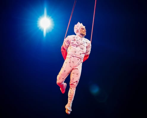A performer in a white costume with black dots, suspended in mid-air by ropes against a dark background with a bright light shining behind.