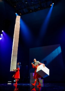 Two performers on stage with dramatic lighting; one balances a tall stack of white boxes while the other holds a single large white box, against a dark backdrop with geometric patterns.