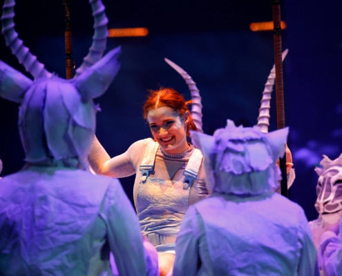 A performer wearing overalls, surrounded by others in horned costumes on stage."