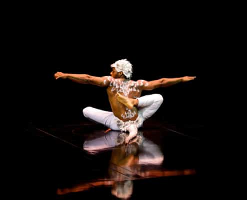 A person with white body paint and headgear, wearing white pants, performing a seated yoga pose with arms extended horizontally on a dark stage.