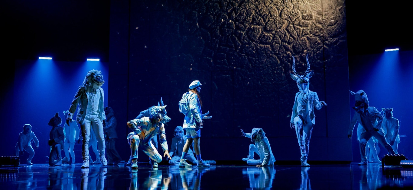 Group of performers in elaborate animal and mythical creature costumes on a blue-lit stage, captured mid-performance with dynamic poses and a spotlighted textured background.