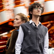 Two people standing in front of a blurred amusement park ride with bright lights and orange structure.