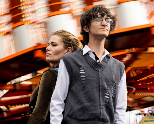 Two people standing in front of a blurred amusement park ride with bright lights and orange structure.