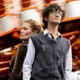 Two people standing in front of a blurred amusement park ride with bright lights and orange structure.