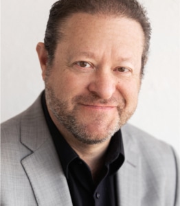 A person wearing a light gray suit jacket over a black shirt, standing against a plain, light-colored background. The man has a close cropped beard and mustache.