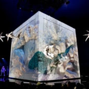 Performers in white costumes suspended in mid-air around a large illuminated cube with nature-themed projections, on a dark stage with blue lighting."
