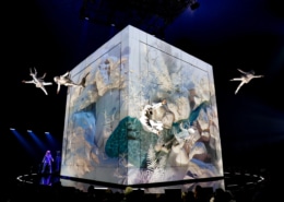 Performers in white costumes suspended in mid-air around a large illuminated cube with nature-themed projections, on a dark stage with blue lighting."