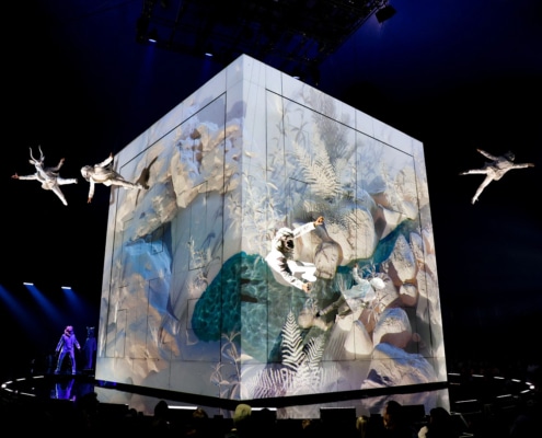 Performers in white costumes suspended in mid-air around a large illuminated cube with nature-themed projections, on a dark stage with blue lighting."
