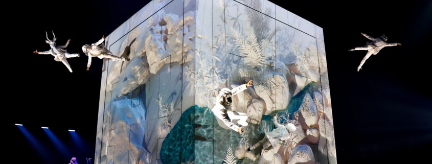 Performers in white costumes suspended in mid-air around a large illuminated cube with nature-themed projections, on a dark stage with blue lighting."