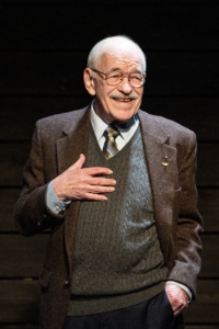 An older man wearing a brown blazer, grey sweater, and tie with one hand on the chest and the other in the pocket.